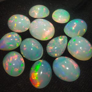 May include: A collection of 12 oval-shaped opal gemstones with iridescent colors, including green, blue, and pink.