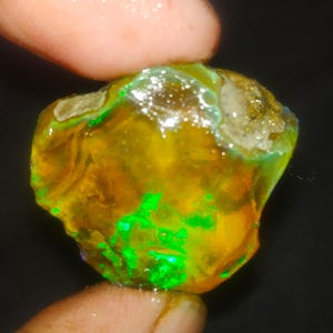 May include: A rough, uncut opal gemstone with a vibrant display of colors. The opal features a mix of yellow, orange, and green hues, with flashes of bright green. The stone is held between two fingers, showcasing its natural, raw form.