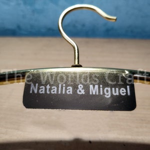 May include: A gold-toned metal clothes hanger featuring a central black rectangular plaque. The plaque is engraved with the white text "Natalia & Miguel" and the words "The worlds Craft" are visible in a faint white font above the name.