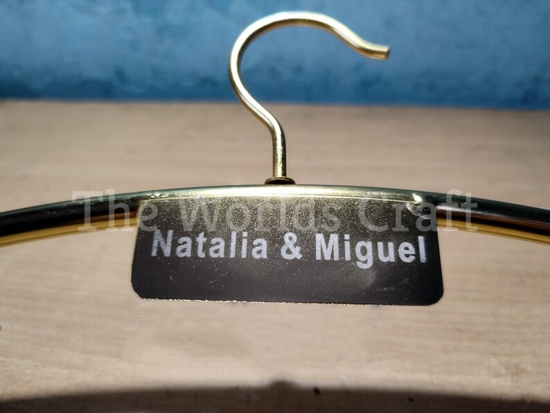 May include: A gold-toned metal clothes hanger featuring a central black rectangular plaque. The plaque is engraved with the white text "Natalia & Miguel" and the words "The worlds Craft" are visible in a faint white font above the name.