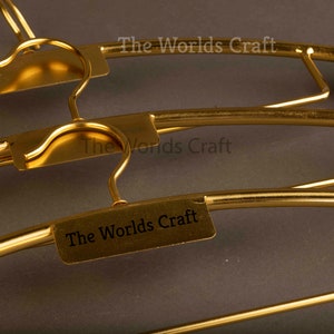 May include: A set of gold-colored metal clothes hangers featuring a curved hook and a rectangular plate attached to the center. Each plate is engraved with the text The Worlds Craft in a clear, dark font, providing a uniform look for the hangers.
