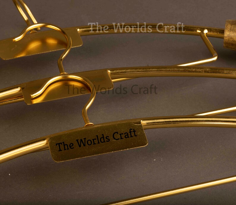 May include: A set of gold-colored metal clothes hangers featuring a curved hook and a rectangular plate attached to the center. Each plate is engraved with the text The Worlds Craft in a clear, dark font, providing a uniform look for the hangers.