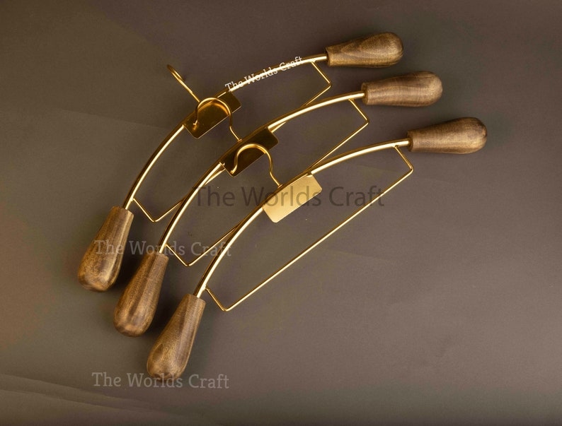 May include: Three clothes hangers featuring gold-toned metal frames and dark brown wooden ends. Each hanger includes a central hook and a rectangular plate with the text The Worlds Craft printed in white. The design combines metallic and wood elements.