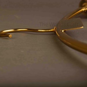 May include: A gold-toned metal clothes hanger featuring a curved hook and a rectangular tag with the text The Worlds Craft printed on it. The hanger has a smooth, polished finish and a curved frame designed for hanging garments in a closet.