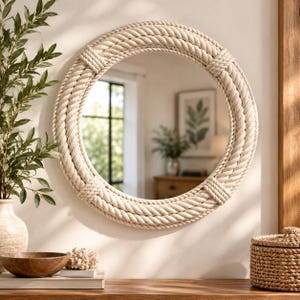 May include: A round wall mirror featuring a thick frame wrapped in natural beige rope. The frame includes four evenly spaced decorative rope knots at the top, bottom, left, and right sides. The mirror surface reflects a bright indoor room.