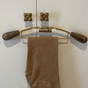 May include: A gold-toned metal clothes hanger with dark brown wooden ends, featuring the text The Worlds Craft printed in white. A pair of folded tan denim jeans hangs over the horizontal bar of the hanger, displaying a single back pocket.