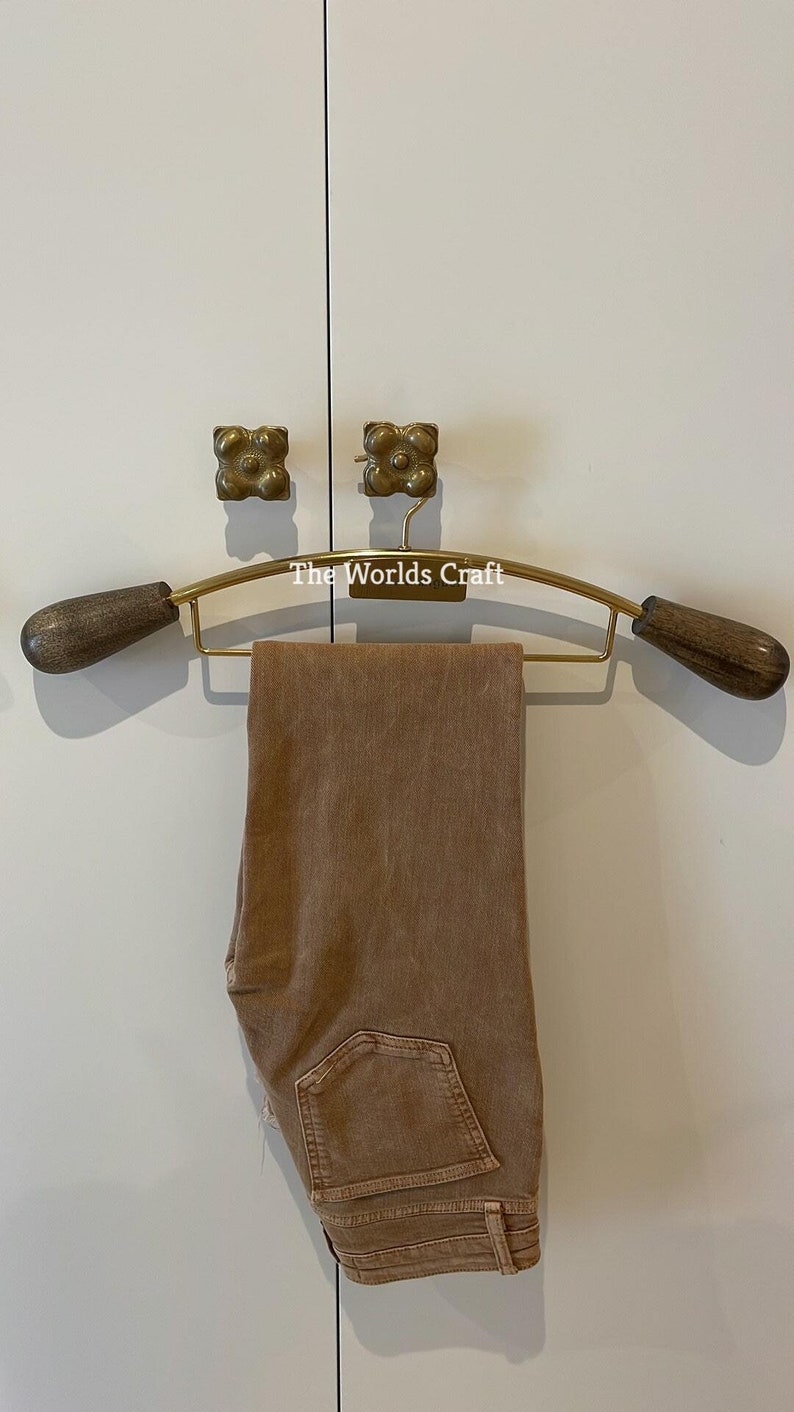 May include: A gold-toned metal clothes hanger with dark brown wooden ends, featuring the text The Worlds Craft printed in white. A pair of folded tan denim jeans hangs over the horizontal bar of the hanger, displaying a single back pocket.