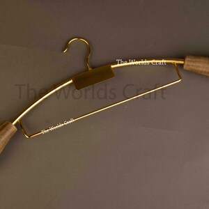 May include: A gold-toned metal clothes hanger featuring a central rectangular plate and two rounded, dark brown wooden end caps. The text The Worlds Craft is printed in white on the metal frame and the central plate of the hanger.