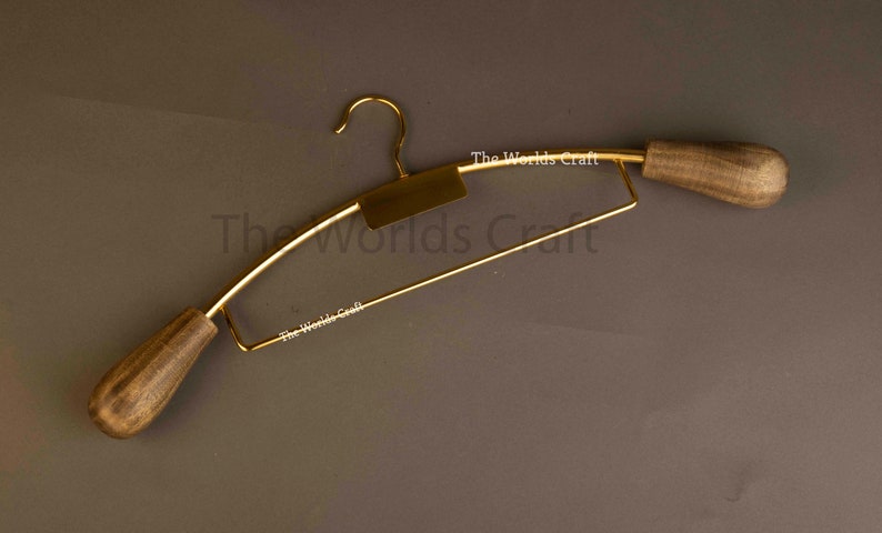 May include: A gold-toned metal clothes hanger featuring a central rectangular plate and two rounded, dark brown wooden end caps. The text The Worlds Craft is printed in white on the metal frame and the central plate of the hanger.