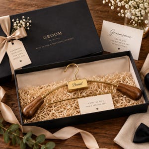 May include: A gold-toned metal hanger with dark wood ends, featuring the name Daniel engraved on a central rectangular plate. The hanger is presented in a black gift box filled with shredded paper, accompanied by a card reading A SPECIAL GIFT.