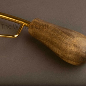 May include: A tool with a polished gold-tone metal frame and a smooth, dark brown wooden handle. The text The World's Craft is visible on the metal surface. The handle has a rounded, tapered shape with a visible wood grain texture throughout.