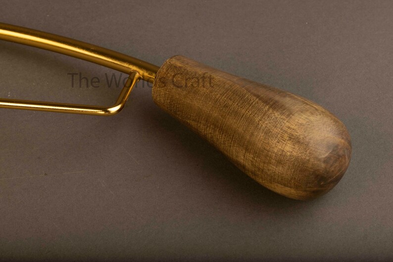 May include: A tool with a polished gold-tone metal frame and a smooth, dark brown wooden handle. The text The World's Craft is visible on the metal surface. The handle has a rounded, tapered shape with a visible wood grain texture throughout.