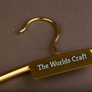 May include: A gold-colored metal clothes hanger featuring a central rectangular plate with the text The Worlds Craft printed in light blue. The hanger has a smooth, polished finish and a curved hook at the top for hanging garments securely.