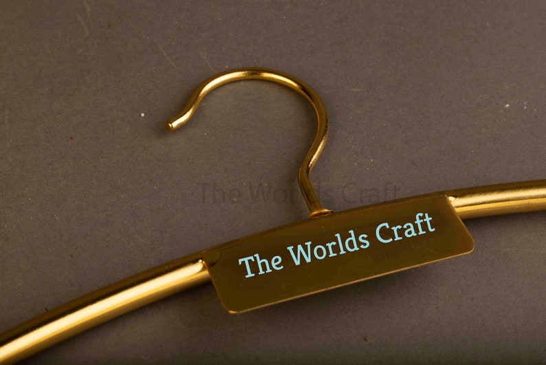 May include: A gold-colored metal clothes hanger featuring a central rectangular plate with the text The Worlds Craft printed in light blue. The hanger has a smooth, polished finish and a curved hook at the top for hanging garments securely.