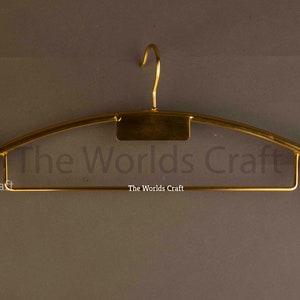 May include: A gold-toned metal clothes hanger featuring a central hook, a rectangular plate, and two rounded, dark brown wooden ends. The text The Worlds Craft is printed on the hanger. The design includes a lower horizontal bar for trousers.