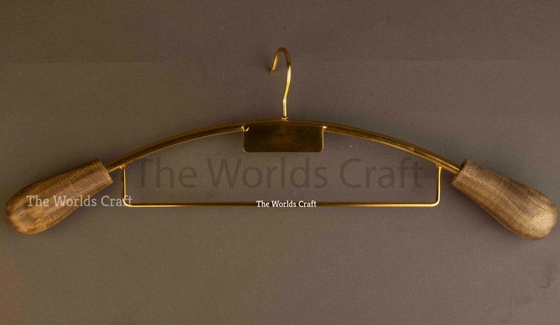 May include: A gold-toned metal clothes hanger featuring a central hook, a rectangular plate, and two rounded, dark brown wooden ends. The text The Worlds Craft is printed on the hanger. The design includes a lower horizontal bar for trousers.