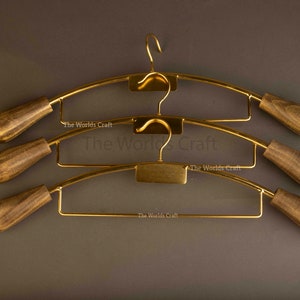 May include: Three gold-toned metal clothes hangers with dark brown wooden ends. Each hanger features a curved top bar, a central hook, and a lower horizontal bar. The text The Worlds Craft is printed on the metal frame of each hanger.
