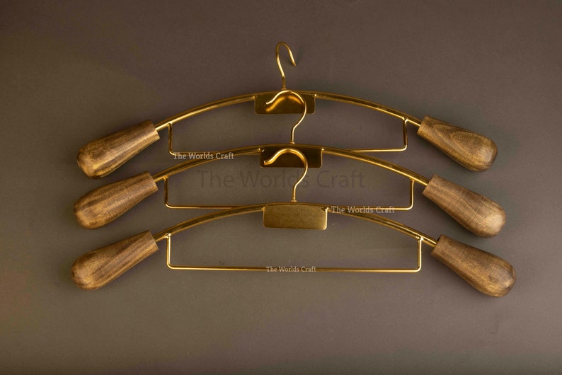 May include: Three gold-toned metal clothes hangers with dark brown wooden ends. Each hanger features a curved top bar, a central hook, and a lower horizontal bar. The text The Worlds Craft is printed on the metal frame of each hanger.