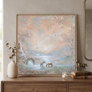 Pode incluir: Uma pintura quadrada emoldurada apresentando uma paisagem texturizada com dois cavalos perto de um pequeno lago, um arco de pedra e uma cachoeira. A cena é renderizada em tons suaves de pêssego, creme e azul claro com três pássaros no céu.
