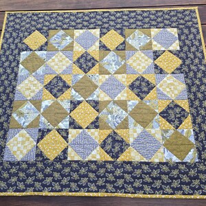 May include: A handmade quilt with a patchwork design. The quilt features a central pattern of diamonds in shades of gold, blue, and brown. A dark blue border with a leaf pattern surrounds the central design, with a gold trim.