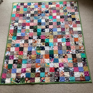 May include: A colourful patchwork quilt with a green border. The quilt is composed of numerous small, square fabric pieces in diverse colours and patterns, including floral, geometric, and text designs. The quilt rests on a neutral-toned carpet.