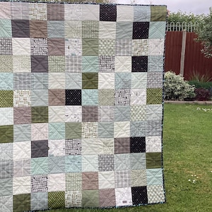 May include: A patchwork quilt featuring a mix of green, brown, and black squares. The quilt is hanging on a washing line in a garden.