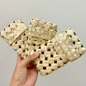 May include: Three woven baskets made of natural materials, with a light brown color and a square shape. The baskets are stacked on top of each other and are held in a hand.