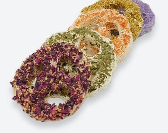 May include: A stack of five circular loofah sponges, each embedded with different dried botanical elements including purple rose petals, green herbs, orange citrus peel, yellow flowers, and a solid purple lavender-colored piece.
