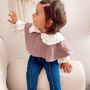 May include: A young child wearing a mauve ruffled bib with a white lace collar, blue jeans, and a pink hair clip. The child is sitting on a white couch.