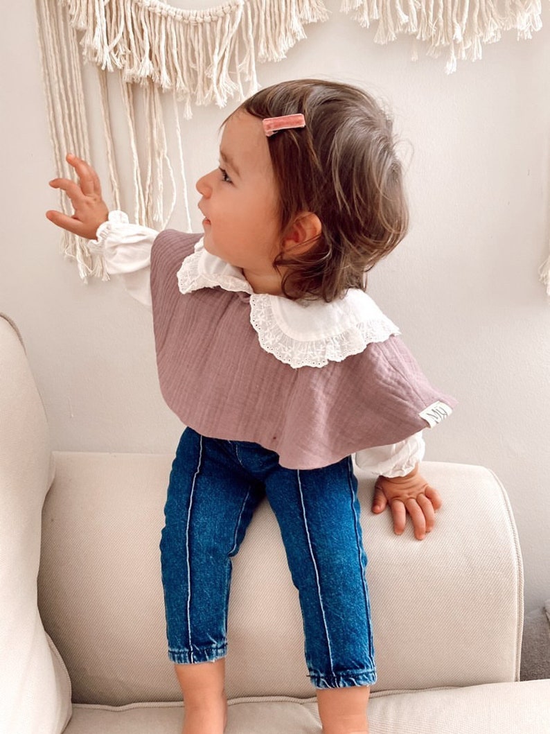 May include: A young child wearing a mauve ruffled bib with a white lace collar, blue jeans, and a pink hair clip. The child is sitting on a white couch.