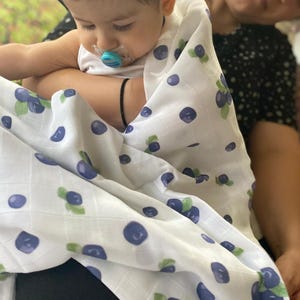 May include: A white baby blanket with a repeating pattern of blueberries and green leaves. The blanket is wrapped around a baby wearing a white onesie and a pacifier.