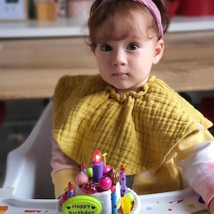May include: A young child wearing a yellow bib and a pink headband sits in a high chair in front of a colorful toy birthday cake with candles. The cake has a pink and white base with a white oval on top that says "Happy Birthday".