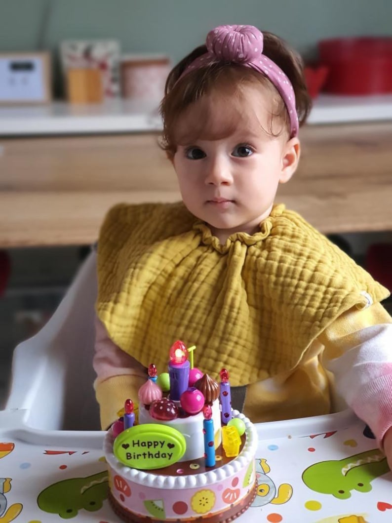 May include: A young child wearing a yellow bib and a pink headband sits in a high chair in front of a colorful toy birthday cake with candles. The cake has a pink and white base with a white oval on top that says "Happy Birthday".