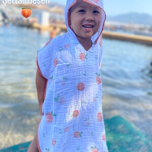 May include: A white hooded poncho made of lightweight muslin fabric featuring a repeating peach pattern with small green leaves. The garment has a peach-colored trim along the hood and edges, with the text "Şeftali desen" and a peach icon.