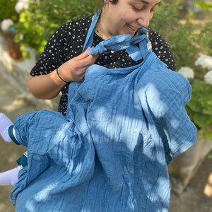 May include: A woman wearing a blue and white patterned shirt holds a blue muslin baby wrap. The wrap is tied around her neck and she is holding the fabric in front of her.