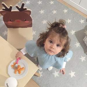 May include: A young child sits at a table with a wooden reindeer chair. The table has a white plate with orange slices and a pink fork. The child is wearing a light blue shirt and has brown hair.