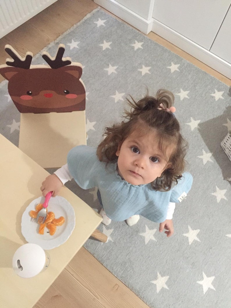 May include: A young child sits at a table with a wooden reindeer chair. The table has a white plate with orange slices and a pink fork. The child is wearing a light blue shirt and has brown hair.