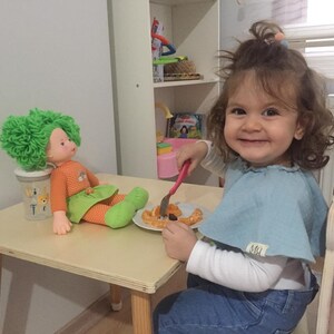 May include: A young girl sits at a wooden table with a doll and a plate of food. The girl is wearing a blue shirt and jeans. The doll has green hair and is wearing an orange shirt and green skirt. The table is light brown and has a white surface. The plate has a fork on it and some food that looks like orange slices and a few small brown pieces.