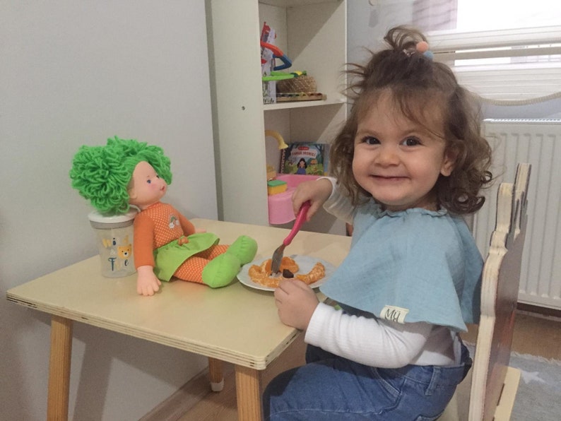 May include: A young girl sits at a wooden table with a doll and a plate of food. The girl is wearing a blue shirt and jeans. The doll has green hair and is wearing an orange shirt and green skirt. The table is light brown and has a white surface. The plate has a fork on it and some food that looks like orange slices and a few small brown pieces.