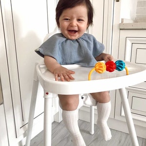 May include: A baby sitting in a white high chair, wearing a light blue shirt and white socks. The baby is smiling and holding a colorful toy.
