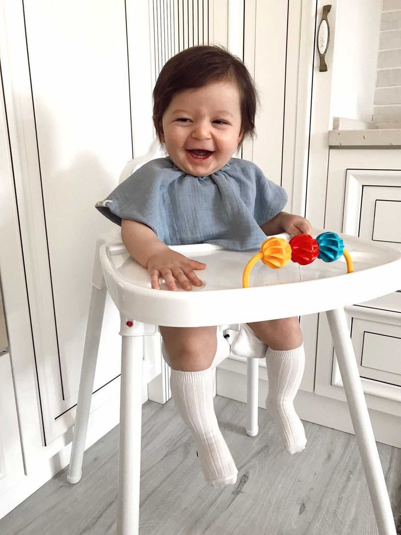 May include: A baby sitting in a white high chair, wearing a light blue shirt and white socks. The baby is smiling and holding a colorful toy.