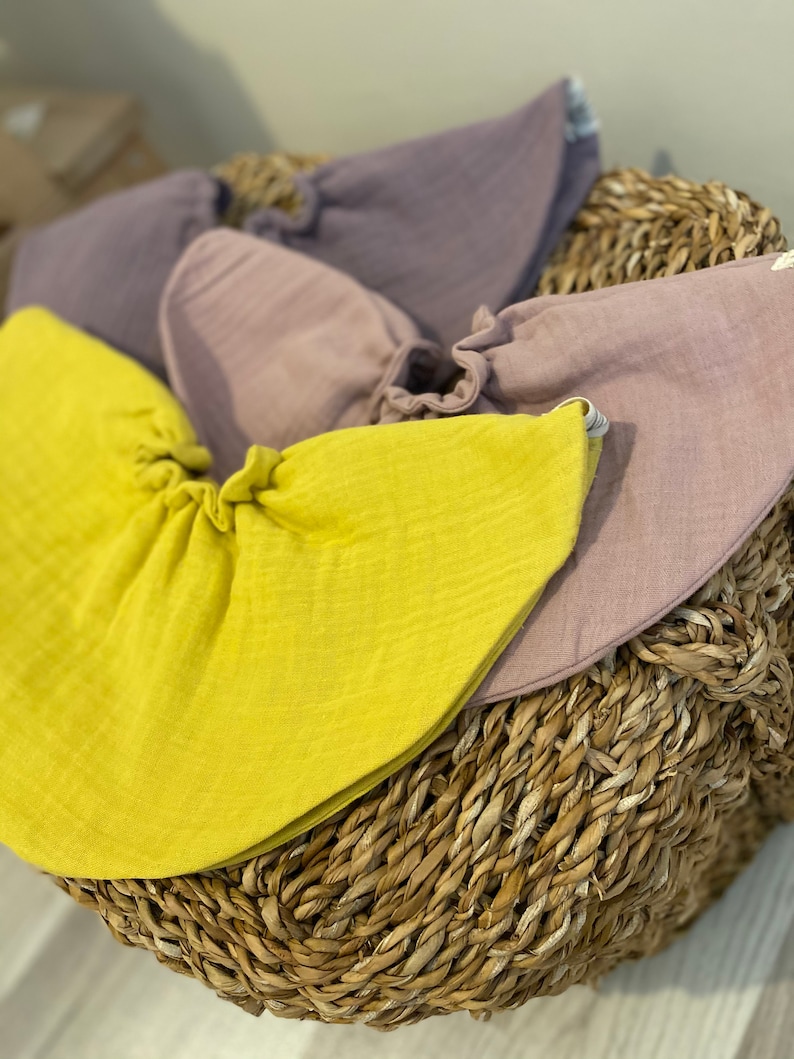 May include: Three muslin baby bibs in a wicker basket. The bibs are in shades of yellow, purple, and light purple.