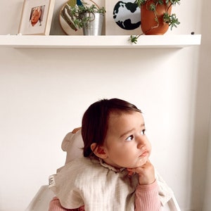 May include: A baby wearing a beige bib with the word "MILK" on it, sitting in a high chair and looking up. The baby is wearing a pink long-sleeved shirt.