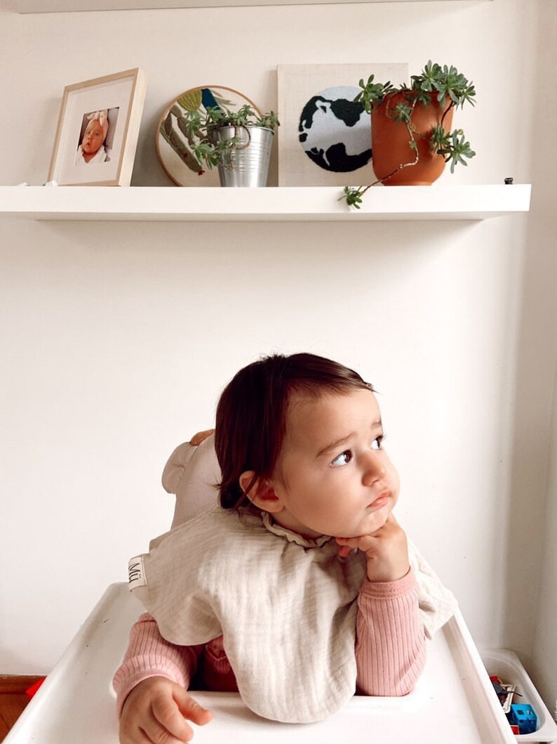 May include: A baby wearing a beige bib with the word "MILK" on it, sitting in a high chair and looking up. The baby is wearing a pink long-sleeved shirt.