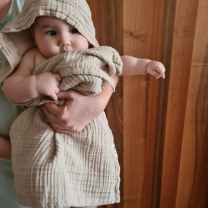 May include: A beige hooded towel with a textured waffle weave pattern. A baby is wrapped in the towel and is looking at the camera.