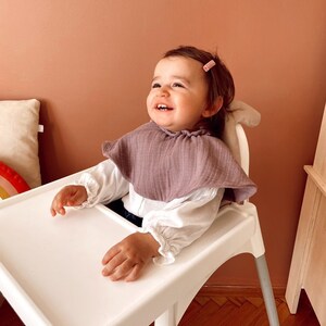 May include: A baby girl wearing a white shirt and a purple bib sits in a white high chair with a white tray. The baby is smiling and looking up. The high chair has a silver frame and is on a wooden floor.