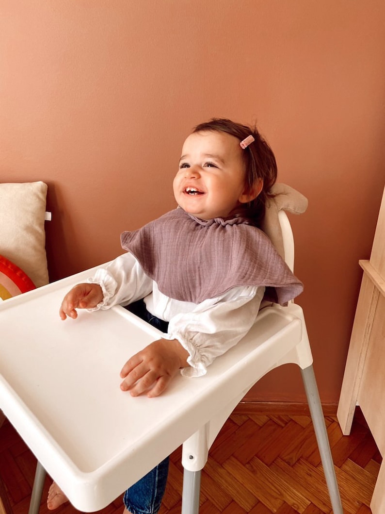 May include: A baby girl wearing a white shirt and a purple bib sits in a white high chair with a white tray. The baby is smiling and looking up. The high chair has a silver frame and is on a wooden floor.
