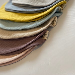 May include: A stack of seven muslin baby bibs in various colors, including beige, light blue, yellow, and pink. The bibs are made of soft, breathable fabric and are perfect for keeping baby's clothes clean.