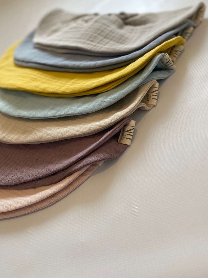 May include: A stack of seven muslin baby bibs in various colors, including beige, light blue, yellow, and pink. The bibs are made of soft, breathable fabric and are perfect for keeping baby's clothes clean.