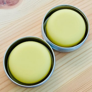 May include: Two round containers with lids, each containing a yellow balm or salve. The containers are made of metal and have a silver finish.